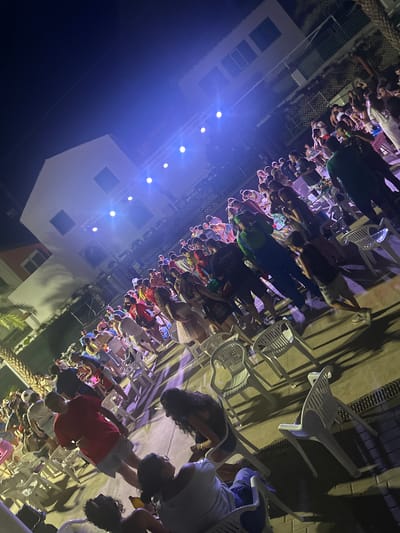 The image depicts an outdoor event at night, likely in Blanca, Spain, given the context. A crowd of people, varying in age and attire, are gathered, with many standing and some seated in white plastic chairs. The scene is illuminated by bright blue stage lights, casting long shadows and creating a vibrant, energetic atmosphere. In the background, a multi-story building with windows and balconies is visible, suggesting an urban or resort setting. Some individuals appear to be dancing or socializing, while others are observing the event. The overall impression is that of a lively outdoor concert or festival.