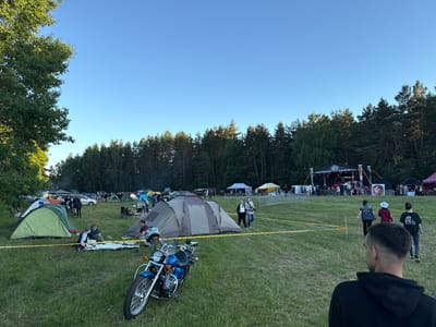 The image depicts an outdoor event, likely a festival or concert, taking place in a grassy field bordered by a dense forest. In the foreground, a blue motorcycle is parked. The middle ground shows a campsite with several tents, people sitting and interacting on blankets, and a yellow caution tape delineating an area. Further back, a stage with lighting and sound equipment is set up, and a crowd is gathered in front of it. Several cars are parked in the background. The sky is clear and blue, indicating daytime. The location is identified as Валожынскі сельскі Савет, Belarus.