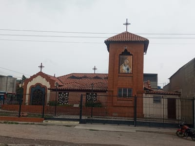 The image shows a church in Barquisimeto, Venezuela. The church is primarily constructed of red brick, with a red tile roof. A tower extends upward from the main structure, featuring a portrait of a religious figure with the words "QUEDATE SENOR CONMIGO" above it. The tower is topped with a cross. The front of the church is surrounded by a black metal fence. A red motorcycle is parked in front of the building. The sky is overcast. The street is unpaved.