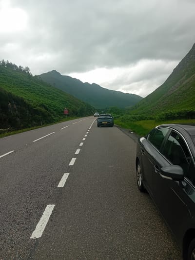 The image shows a road in a mountainous, green landscape, likely the A82 near Invergarry, United Kingdom. The road is paved and has white dashed lines marking the lanes. There are several cars on the road. In the foreground, a dark-colored car is parked on the right side of the road, with its rear end visible. Further down the road, two other cars are driving, a dark SUV in the middle lane and a lighter-colored car in the left lane. The landscape is characterized by steep, green hills covered in vegetation and trees, with a cloudy sky overhead. A red sign with a warning symbol is visible on the left side of the road. The overall impression is of a scenic drive through a remote, natural area.