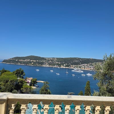 The image captures a panoramic view of the bay in Saint-Jean-Cap-Ferrat, France, from a vantage point behind an ornate balcony railing. The sky is a clear, vibrant blue, stretching across the upper portion of the frame. The body of water is a deep, rich blue, dotted with various white vessels, including several large yachts and smaller boats. Along the coastline, a lush green hillside is densely populated with buildings, characterized by their terracotta roofs and pastel facades. Some of these structures appear to be villas nestled amongst the trees, while others form a more concentrated townscape along the water's edge. The vegetation is abundant, with dark green cypress trees and olive trees framing the scene, particularly on the right side of the image. In the foreground, the decorative white stone railing of a balcony, featuring intricate carvings and pillars, partially obstructs the view, adding a sense of depth and perspective. The overall impression is one of a serene, upscale coastal landscape on a bright, sunny day.