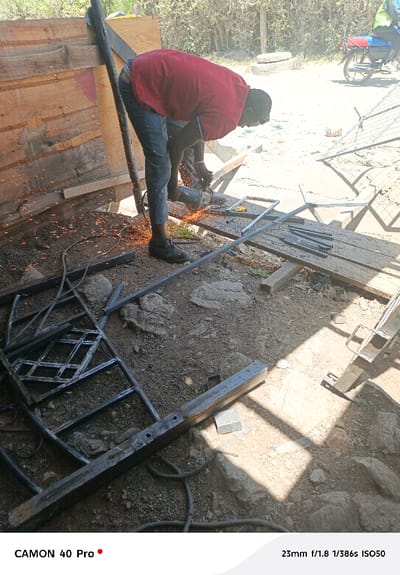 A person in a red shirt and jeans is using a grinder to cut metal. Sparks are flying from the grinder as it cuts through the metal, with a partially completed metal structure lying on the ground, along with other cut pieces of metal. The ground is covered in dirt, small rocks, and metal debris. A wooden structure is visible in the background, and a motorcycle is parked further away.