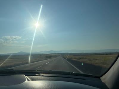 The image captures a view from inside a car, looking out through the windshield. The sun is high and bright in a clear blue sky, creating lens flare and streaks of light across the windshield. The windshield itself has a prominent crack. The car appears to be on a road, with a flat, grassy landscape stretching out on either side. In the distance, there are rolling hills or mountains under a hazy sky. The foreground shows the dashboard of the car, with its textured surface. The scene suggests a journey on a sunny day, likely on a highway outside of a populated area. The specific location in Yeghvard, Armenia is not visually identifiable from the image itself.