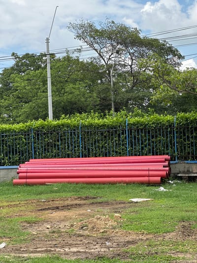 In the city of Juan Mina, Colombia, a stack of bright red cylindrical pipes lies horizontally on the ground in a grassy area. The pipes are arranged in multiple layers, with the uppermost layer positioned slightly forward, creating a staggered effect. Behind the pipes, a black metal fence with blue vertical posts runs across the scene, topped with a thick hedge of lush green foliage. A concrete wall with some visible bricks is situated to the right of the pipes, next to the fence. In the background, a variety of trees with vibrant green leaves form a dense canopy, obscuring the horizon. Power lines are visible crisscrossing the upper portion of the image, and a tall, light-colored utility pole with an angled arm extends upwards into the cloudy sky. The ground in front of the pipes is a mix of green grass and patches of exposed brown soil, with some tire tracks visible in the dirt. The overall environment suggests an outdoor, possibly construction or storage area.
