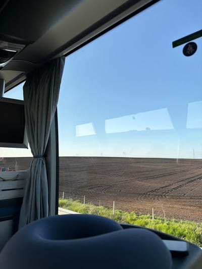 The image, seemingly taken from the interior of a vehicle, shows a landscape adjacent to a field. The vehicle's window offers a view of a cultivated agricultural field, characterized by its dark soil and distinct rows. A strip of green vegetation borders the field. Utility poles with power lines are visible in the distance, stretching across the field. The sky is a clear blue, with the sun seemingly high overhead. Inside the vehicle, grey curtains are partially drawn, and a blue headrest is also visible. The interior is visible with vehicle's trim and control panels. The city of Zetiovo, Bulgaria is not confirmed in this image, but it is a possible location.