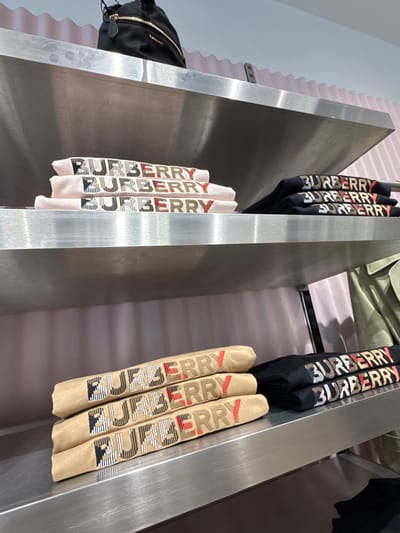 The image depicts a retail display at a Burberry store located in La Roca Village, La Roca del Vallès, Spain. On the top shelf, a black Burberry bag with gold hardware is visible. Below, on the same shelf, are stacks of folded light pink t-shirts, each emblazoned with the Burberry logo in various checkered patterns. To the right, on the same shelf, are stacks of folded black t-shirts, also featuring the Burberry logo with checkered detailing. The lower shelf displays a stack of three tan-colored rolled t-shirts, each with the Burberry logo embroidered in a checkered pattern. Next to these rolled t-shirts are two folded black t-shirts, again with the Burberry logo. A beige trench coat is partially visible hanging on the right side of the lower shelf. The background consists of a pale pink wall with a corrugated texture. The display shelves are made of brushed metal.