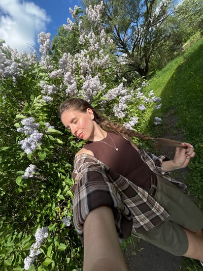 A woman with light skin and brown braided hair is lying amidst a bush of lilac flowers in the city of Борщевка Малая, Russia. She is wearing a brown tank top, olive green pants, and a brown and beige plaid shirt. The woman is holding the end of her braid in her right hand. In the background are a tree, grass, and a path. The sky with white clouds is visible in the upper left corner.