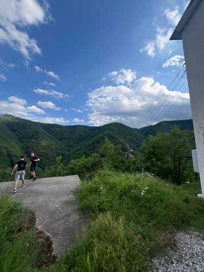 The image depicts two young males standing on a concrete surface in an outdoor, hilly, and wooded environment. The male on the left is wearing a black t-shirt and light-colored shorts. The male on the right is wearing a black t-shirt and black shorts. They are facing towards a lush green valley with densely forested hills. The sky is blue with scattered white clouds. To the right of the scene, a portion of a white building with a sloped roof is visible, along with power lines. The concrete surface appears to be part of an elevated structure, possibly a platform or the edge of a road. Vegetation, including tall grass and small white flowers, grows around the concrete. The overall impression is a scenic view from a relatively high vantage point overlooking a natural landscape. The image does not provide specific location details within the city of Pechinska, Bulgaria.