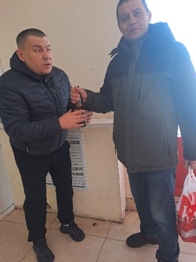 Two adult males are depicted standing on a tiled outdoor surface in Bolshaya Elhovka, Russia. The male on the left, wearing a dark blue puffer jacket, black pants, and dark athletic shoes, holds a dark glass bottle with both hands and appears to be speaking, looking towards the right. The male on the right, dressed in a dark olive green and black winter jacket, blue jeans, and dark shoes, is holding the same dark glass bottle with his left hand, interacting with the first male. He also carries a red and white plastic shopping bag in his right hand. The background features a plain, light-colored wall with a lower ledge and a white horizontal slatted structure. The floor consists of light-colored square tiles.