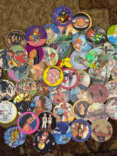 This media file is a top-down, close-up shot of a collection of round, collectible game pieces, often referred to as "pogs." The pogs are scattered on a textured surface, likely a rug or carpet, which appears to be a brownish-orange color, suggesting an indoor setting. The lighting is diffused, casting soft shadows and highlighting the glossy surfaces of many of the pogs, some of which have a holographic sheen.
The subjects depicted on the pogs are varied, featuring cartoon characters, animated figures, and what appear to be illustrations of people in action, some of whom are engaged in sports like basketball or soccer. There are also pogs with depictions of animals, possibly fantastical creatures, and some with images of people's faces. One pog prominently features a drawing of a character with a yellow head and large, expressive eyes, while another shows a woman reclining. The style of the artwork ranges from simplistic cartoon drawings to more detailed illustrations.
Visible text on the pogs includes brand names and copyright information, such as "POWER RANGERS," "FLIPCAP," "UNIVERSAL," "SHOCK," "ROCKSTEADY," "METEOROPOG," "SAUS POGA," and "TM & © 1995 WPF." There is also some text in what appears to be Cyrillic script, including "ПЛАНЕТА ОБЕЗ," and "ALROG." The presence of these names and copyright dates suggests the pogs are from various entertainment franchises and were produced around the mid-1990s. The overall impression is a collection of colorful and diverse game pieces, indicative of a hobby or childhood pastime. There are no clear indications of a specific time of day, weather conditions, or emotions, as the focus is on the objects themselves. The location context provided, Rzhev, Russia, is not directly inferable from the visual elements of the image.