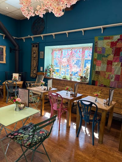 The image shows the interior of a room with colorful and eclectic decor. The walls are painted a deep teal, and a window is adorned with pink cherry blossoms, letting in natural light. Above the window, a patchwork quilt hangs. Tables of various wooden designs are scattered around the room, some set with flowers in small vases and menus. Chairs of different colors and styles, including blue, pink, and a floral-patterned green folding chair, are arranged around the tables. A black stove pipe is visible against the wall, and decorative items such as a circular metal wall hanging and a framed chalkboard add to the room's character. The location of the scene is in the city of West Oxfordshire, United Kingdom.