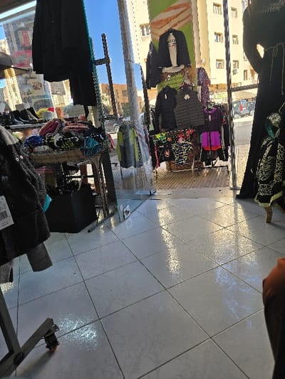 The image captures the interior of a clothing store in Taza, Morocco, observed from a low vantage point looking towards a large glass storefront. The floor consists of light-colored, square tiles that exhibit reflections of exterior light sources. In the foreground, the lower portion of a human leg, dressed in dark fabric, is visible on the right.

The midground features various merchandise displays. To the left, a display table holds an assortment of black boots, colorful knitted headwear, and other accessories. A black bag is positioned beneath this table. Multiple metallic clothing racks are arranged, some visible directly inside and others through the glass storefront. These racks display a range of garments, including black puffer jackets, a grey puffer vest, a white t-shirt on a display bust, and various colorful shirts and dresses. A brown woven basket is situated beneath one of the central display racks.

Through the glass storefront, the exterior environment is visible under clear, bright conditions. A light beige, multi-story building with numerous windows stands across the street. A vehicle, possibly a car or van, is present on the street. A clear blue sky is visible above the buildings, and a green and orange striped banner or sign is partially visible above the storefront's exterior section.