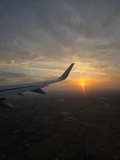 The image shows a sunset view from an airplane window. The right wing of the plane, with the Wizzair logo, is visible in the foreground. The sun is positioned on the horizon, emitting bright rays. Below the plane, the ground appears to be composed of fields. The environment appears to be in a rural area. The location is in Dacorum, United Kingdom.