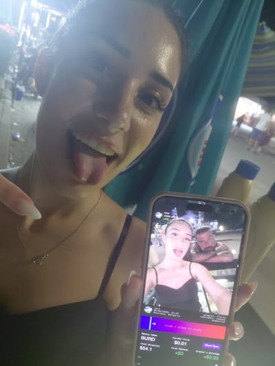 The image captures a close-up selfie taken by a woman in Kostievo, Bulgaria, at 21:47, as indicated on the displayed phone screen. The woman in the foreground is smiling broadly with her tongue extended and is pointing towards her chin with her right index finger, which has a long, pointed, light-colored nail. She wears a dark, thin-strapped top and a delicate silver-colored necklace with a small heart-shaped charm. She holds a smartphone in her left hand, displaying a screen that shows another selfie.
The smartphone screen features an image of a woman, resembling the foreground subject, also with her tongue extended, alongside a bearded man with visible arm tattoos, who is leaning forward with his arms crossed. Below this image, text states "vivi," "05/05/2026, 21:47," and "Kostievo, Bulgaria," along with descriptive terms: "playful, flirty, confident, cheeky, seductive." Additional information on the screen details a "Memory Token BURD" with a "Current Price $0.01," "Total Attention $54.1," "Total Revenue +$0," and "Creator's Earnings +$0.55," accompanied by a "Swipe / click to stake" prompt.
The background behind the foreground woman is an outdoor, nighttime scene. A prominent vertical section of blue-green fabric, possibly an awning or banner, is visible on the right side. To the right of her head, two yellow bottles with blue caps are partially discernible. The distant background is out of focus, revealing blurred figures of people and multiple bright light sources, suggesting a lively street or public area.