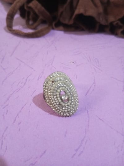 An elongated, oval-shaped ring is positioned on a light purple, textured surface in Chandragiri, Nepal. The ring's decorative face features concentric oval patterns composed of small, light-colored spherical beads, interspersed with rows of clear, faceted stones. A larger single clear stone is embedded near the approximate center of the design. The ring casts a distinct shadow to its lower-left. In the upper portion of the frame, a crumpled mass of dark brown fabric is visible out of focus. No individuals or direct interactions are present in the scene.