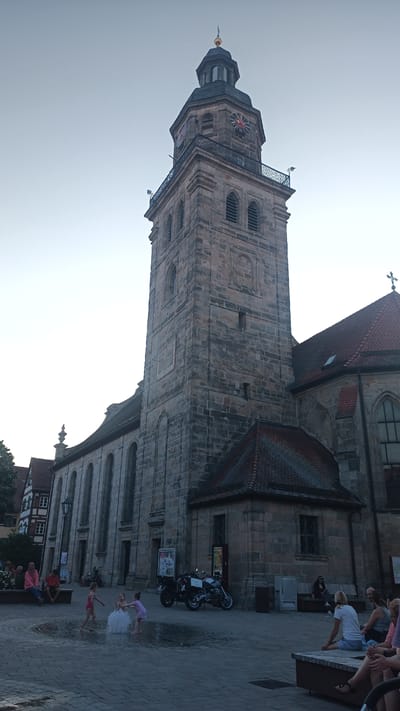 The image depicts a low-angle view of a large stone church with a prominent bell tower. The tower features multiple clock faces with red numerals and hands, and arched windows. A sundial is also visible on one side of the tower. The church has a red-tiled roof and a cross at its apex. In the foreground, a paved area with a water fountain is the setting for children playing. One child in a white dress is surrounded by splashing water, while two other children in swimwear play in the fountain. Several adults are seated on benches in the background, some observing the children. Two motorcycles are parked near the church entrance. The scene appears to be outdoors in a town square. The lighting suggests it is late afternoon or early evening, with a soft, diffused light. The sky is clear and pale. The architectural style of the church and the adjacent buildings, along with the German text on posters displayed on the church wall, suggest the location is Altdorf bei Nürnberg, Germany. The overall atmosphere is relaxed and pleasant, with families enjoying a public space.