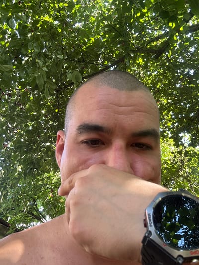 A person is taking a selfie outdoors, with their head and shoulders filling most of the frame. They have a shaved head and are wearing a watch on their left wrist. Their hand is raised, covering their mouth. The background is a canopy of green leaves with some red berries, suggesting they are under a tree. Sunlight filters through the leaves, creating a dappled effect. The scene is set in the city of Velykyi Trostianets, Ukraine.