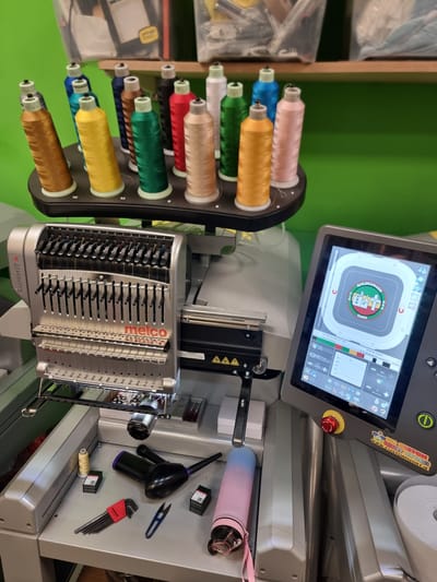 This media file shows a close-up, rotated view of an industrial embroidery machine, likely in a workshop or studio in Tullamore, Ireland. The machine, identified as a Melco Summit, is the primary subject.
The machine features a prominent touchscreen display showing a complex embroidery design of a town crest with the words "RAILENAMAOR DARU" and some Irish text. The display also shows various operational parameters like stitching progress, time remaining, and stitch count. In front of the display is a red emergency stop button.
To the left of the machine, a rotating spool holder is filled with spools of colorful embroidery thread, including pink, blue, orange, green, red, and gold. These threads are feeding into the embroidery head of the machine.
The embroidery head itself is a complex metallic component with numerous needles and thread guides, indicating its capability for multi-color embroidery.
Various tools and accessories are visible on the machine's surfaces. These include a pink and blue gradient-colored tumbler, a small pair of scissors, a can of compressed air or a blower, a set of hex keys, and small boxes that likely contain bobbins or other sewing supplies. The "THE CUSTOM SHIRT STORE www.CUSTOMSHIRT.IE" sticker on the machine suggests it is used for commercial garment customization.
The background is a vibrant green wall, typical of a workshop or retail environment. The lighting appears to be artificial and bright, suggesting the image was taken during operational hours. No people are visible in the image. The overall scene conveys a sense of focused activity and the technical nature of the embroidery process.