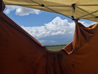 The image presents a view of the Armenian landscape through a frame created by draped brown fabric, likely part of a temporary shelter or tent, with a beige-colored canopy overhead. The canopy is supported by metal poles, and the fabric is tied at one of the supporting poles. The landscape in view consists of green fields leading to a body of water in the distance, beyond which lies land under a sky filled with large, cumulus clouds and patches of blue. Based on the information provided, it is inferred that this scene is set in Tlik, Armenia.
