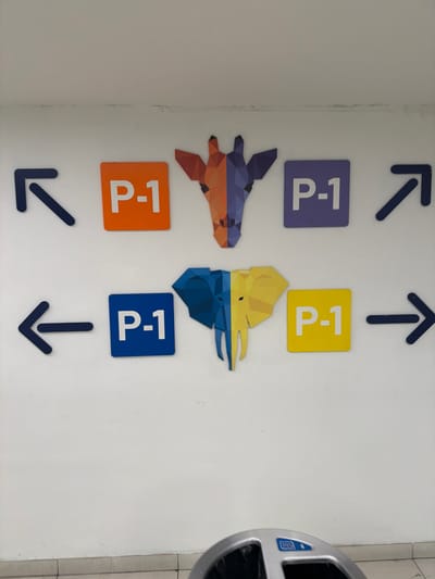 The image shows a white wall with several decals and a trash receptacle. There are two sets of directional arrows, one pointing upwards and to the left, and another pointing upwards and to the right. Below these arrows are two square decals, one purple and one yellow, both displaying "P-1" in white text. Further down, there are two more square decals, one orange and one blue, also with "P-1" in white text. Between the two sets of "P-1" decals are two abstract, geometric animal figures. The figure on the left appears to be a bird or reptile with orange and purple coloring, while the figure on the right is a bird with yellow and blue coloring. Below the lower "P-1" decals are two more directional arrows, one pointing down and to the left, and the other pointing straight down. To the right of the wall, a portion of a silver trash receptacle with a blue logo is visible. The lighting suggests an indoor environment. There is no indication of people, weather, or specific time of day. The location context is Alcorcón, Spain.
