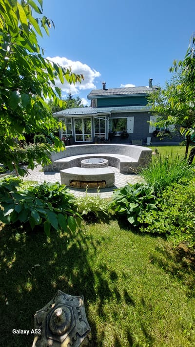 The image depicts a backyard scene in the city of Zhavoronki, Russia, on a sunny day with a bright blue sky and scattered clouds. In the foreground, lush green grass covers the ground, with shadows cast by trees. A decorative stone object with an ornate pattern sits on the grass on the left side.
Dominating the mid-ground is a circular stone fire pit area with a stone bench built around it. The fire pit itself is metal with a grate. Small logs are stacked neatly on a stone surface within the fire pit structure. The bench is constructed from large, flat stones.
Behind the fire pit area is a house with a green roof and grey brick siding. A glass-enclosed sunroom extends from the house, featuring large windows and doors that are partially open. Flower pots with blooming flowers are visible on the ledge of the sunroom and on the ground nearby.
The environment is that of a well-maintained backyard, suggesting a residential property. There are no people or animals visible in the image. The overall scene conveys a sense of outdoor leisure and relaxation.