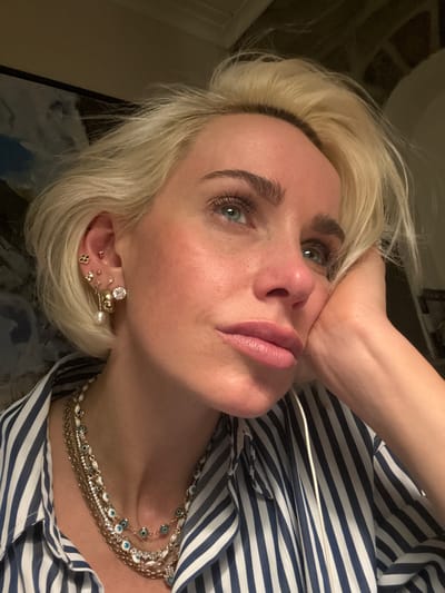 A portrait of a blonde woman wearing a blue and white striped shirt, multiple gold necklaces, and a variety of earrings. She is looking up at the camera with her hand near her face. The location in Strömstad, Sweden, is unknown based on the provided image.