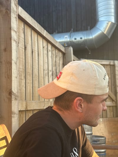 The image depicts the right profile of a seated adult male in an outdoor setting. He wears a light beige corduroy baseball cap with a peach-colored brim and a dark, possibly black, short-sleeved top with a partial white graphic. His short hair is visible beneath the cap, and his face is clean-shaven. The background features a light-colored wooden fence or partition, constructed from vertical planks and horizontal support beams. Behind the fence, a dark, likely black, vertical wall or building exterior is visible. A large, silver-colored metallic ventilation duct system, featuring a distinct curve and visible fasteners, is mounted against this dark wall. In the lower right of the frame, two silver metallic beer kegs are stacked vertically. A portion of a light-colored wooden chair is visible on the far left. The scene is illuminated by warm, golden light, suggesting late afternoon or early evening. The environment is consistent with an outdoor seating area of a commercial establishment, such as a pub garden or brewery taproom, within Northallerton, United Kingdom.