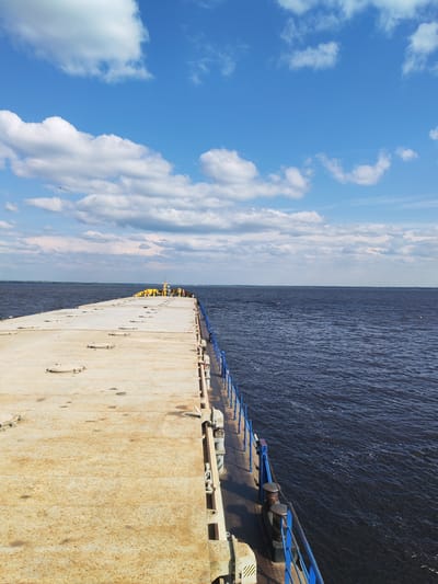 The image depicts a view from the deck of a barge or vessel, looking forward and slightly to the right. The deck is made of a weathered, light-colored material, possibly metal, with visible circular hatches or covers. A blue metal railing runs along the right side of the deck. The dark, rippling water of a wide river or lake extends to the horizon. The sky is bright blue with scattered white cumulus clouds. In the distance, along the horizon, a strip of land is visible, with some structures or lights appearing on the far right. A few yellow objects are clustered on the deck in the distance. The location is identified as Veregonets, Russia.