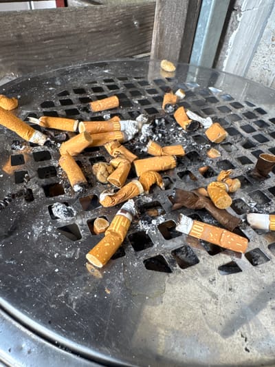 The image shows a close-up of a metal ashtray filled with numerous cigarette butts. The ashtray appears to be outdoors, possibly on a public bench or table, given the presence of what looks like wooden slats in the background, suggesting an urban or park setting. The cigarette butts are varied in their state of consumption, with some appearing freshly discarded and others burnt down. Ash and small debris are scattered around the ashtray's surface. The overall impression is one of a well-used public amenity. The image does not provide specific information about the city of Sankt Wendel, Germany.