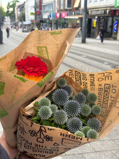 The image displays two bouquets of flowers wrapped in brown paper, held by a person's hands in the foreground. The bouquet on the left contains a bright red Gerbera daisy and an orange flower beneath it. The bouquet on the right features numerous spherical, blue-green flowers with spiky petals. Both bouquets are wrapped in brown paper with green lettering and decorative motifs. In the background, a blurred city street scene is visible, with buildings, shops, and a few pedestrians. The presence of tram tracks on the street indicates an urban environment. The shops have illuminated signs. One sign reads "KREMHANATIV". The overall atmosphere suggests a casual, outdoor setting in a German city. The style of the shops and the German text on the wrapping paper suggest the location is likely Witten, Germany.