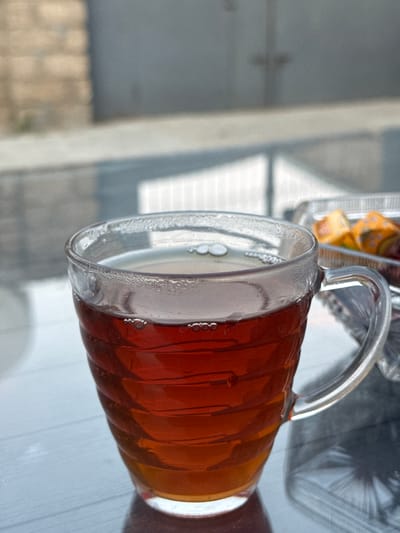 A close-up, angled shot shows a clear glass mug filled with amber-colored tea. The mug has a ridged texture and a handle. Small bubbles are visible in the tea and on the inside of the glass, suggesting it might be hot or carbonated. The mug rests on a reflective surface, possibly a table, which shows a distorted reflection of the mug and its contents. In the background, out of focus, there is a hint of a metallic grate and a light-colored wall or surface. To the right of the mug, a small container with what appear to be individually wrapped candies, possibly with yellow and orange wrappers, is partially visible. The lighting suggests it is daytime, and the overall scene feels like it could be an outdoor setting, perhaps a cafe or a patio, given the background elements. No people are visible in the frame. The location context is Khazar Raion, Azerbaijan.