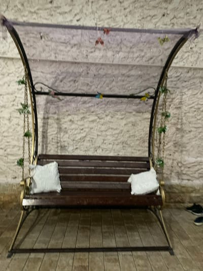 This media file depicts a tranquil outdoor scene in Khasavyurt, Russia. The focal point is a dark wooden bench adorned with two white square cushions, inviting relaxation. The bench is framed by a black metal structure with curved elements, hinting at a decorative garden feature or a small gazebo. Delicate green foliage is interspersed along the metalwork, adding a touch of natural beauty to the man-made structure. Small, colorful bird-like ornaments are attached to the central vertical support of the metal frame. The background consists of a textured, light-colored wall, possibly made of stone or stucco, with a subtle shimmering effect from scattered lights. To the right, a wooden planked surface creates a contrasting texture, suggesting a patio or pathway. In the upper right corner, the feet of two individuals are visible, wearing dark and blue shoes, implying their presence nearby. The lighting suggests it might be evening or a dimly lit area, casting soft shadows and highlighting the reflective elements. The overall atmosphere is one of peaceful serenity.