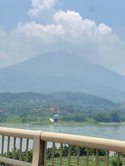 The image shows a hazy mountain range in the distance, with a body of water in the foreground. A tan-colored railing runs across the bottom of the frame, obscuring a portion of the view. Behind the railing, banana trees and what appear to be agricultural fields are visible. Across the water, lush green hills dotted with trees and some structures lead up to the misty mountains. A white cylindrical building, possibly a water tower, is situated on the far bank of the river. The sky is blue with scattered white clouds. The overall scene suggests a rural or semi-rural landscape in Tu Vũ, Vietnam.
