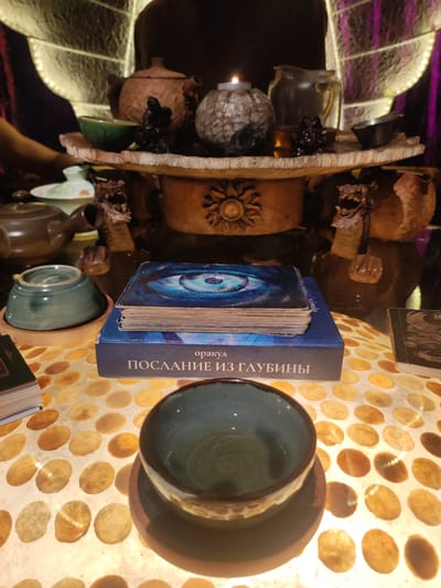 The image depicts an altar-like table, likely indoors, with various objects associated with spiritual or divinatory practices. A stack of cards with a blue eye design on top rests on a box labeled "ПОСЛАНИЕ ИЗ ГЛУБИНЫ". A teal-colored cup sits on the table in the foreground. Another cup is resting on a saucer, stacked on top of another cup, to the left of the cards. The table has a patterned surface with what appears to be round, light-colored spots. A shelf above the table holds teapots, a candle, and other small vessels. The shelf support columns are adorned with carved dragon heads. The overall setting is dimly lit and appears somewhat mystical. The location is not discernible from the image content.