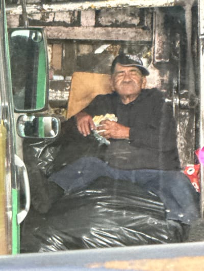 An elderly man is seated on a black plastic bag, likely trash, with a light brown chair behind him. He is wearing a dark long-sleeved shirt and blue pants, and a dark baseball cap. The man holds a sandwich in his right hand and appears to be eating it. His left hand is resting on his lap. The background is out of focus, but appears to be an urban environment with exposed brickwork and metal structures. The scene suggests a setting in Tizapan, Mexico, potentially a street or an alleyway where refuse is stored. A green object, possibly a part of a vehicle or structure, is visible to the left of the man.