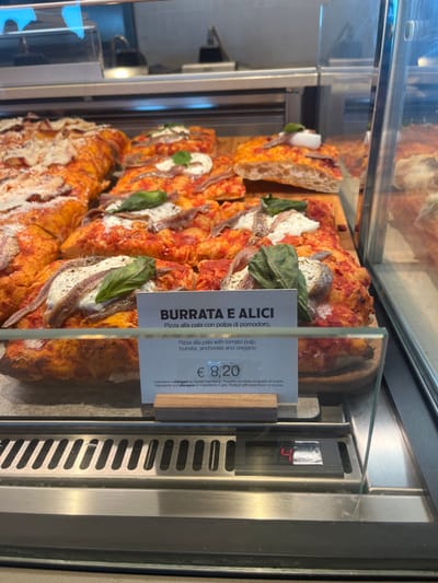 The image captures a close-up of a glass display case showcasing several slices of "Pizza alla pala" (paddle pizza) in a food establishment in Dorno, Italy. The pizzas are rectangular with a thick, airy crust, and are arranged on a wooden-textured surface.
The most prominent pizza slices are generously topped with a rich red tomato pulp, creamy white burrata cheese, distinctive silvery anchovy fillets, and fresh green basil leaves. Other slices appear similar, some featuring the cheese and tomato base.
A clear plastic price tag or menu sign stands upright in front of the pizzas. The main heading reads "BURRATA E ALICI" (Burrata and Anchovies), and the description details the ingredients in both Italian and English: "Pizza alla pala con polpa di pomodoro, burrata, acciughe e origano" (Pizza alla pala with tomato pulp, burrata, anchovies and oregano). The price is clearly displayed as "€ 8,20". Smaller text on the tag indicates "Ingredienti e allergeni" (Ingredients and allergens) and notes that the base is "Prodotto con base congelata all'origine" (Product with base frozen at origin).
The display case features glass panels and stainless steel elements, including a grated vent on the right side. A small digital display, partially visible on the right, shows the number "4." The scene suggests an indoor commercial setting, likely a deli, bakery, or cafeteria, where food is presented for sale.