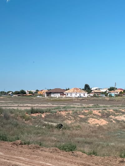 The image depicts a rural landscape with a clear, bright blue sky above. In the mid-ground, a collection of houses and buildings is visible, some appearing to be under construction. One house prominently features a wooden roof structure, indicating ongoing building work. A white car is parked near the houses. The foreground consists of a dry, grassy field with patches of exposed earth and a culvert or pipe running through the vegetation. The overall impression is of a developing or sparsely populated area on the outskirts of a settlement.