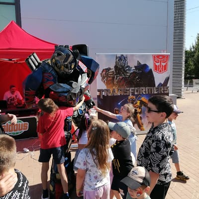 This image depicts an outdoor event in Трубачеевка, Russia, under clear skies. The central focus is a large, detailed costume resembling Optimus Prime from the Transformers franchise, primarily colored blue, red, and grey, featuring shoulder-mounted pipes. Several children are gathered around the costume, engaging with it; one boy in a red shirt and dark shorts is touching its arm, and a girl in a light blue top with sunglasses is reaching towards it.
To the left, under a red pop-up tent, an adult male with a mustache is operating DJ equipment, including a laptop and a mixer, positioned on a table. A sign with the word "GLODUS" in green and orange lettering, accompanied by a globe icon, is visible below the DJ setup. White cables are visible on the reddish-brown paved ground in front of the tent.
In the background, a large vertical banner displays images of Transformers characters, specifically Optimus Prime and Bumblebee, along with the "TRANSFORMERS" logo and the red Autobot symbol. Behind the banner and extending further right is a light-colored building with a distinctive facade featuring horizontal slats. Green trees are visible beyond the building on the right, and a metal shopping cart is partially visible in the far right background. The ground throughout the visible area is paved with interlocking bricks.
