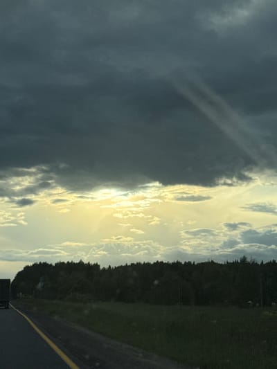 The image depicts a dramatic sky over a natural landscape. The sky is a prominent subject, characterized by a stark contrast between dark, heavy clouds on the left and bright, sunlit clouds on the right. Rays of sunlight pierce through the brighter clouds, creating a luminous and ethereal effect. This suggests a transitional weather condition, possibly a storm moving in or passing by, with the sun attempting to break through.
The scene below the sky includes a dark line of trees, likely a forest, running horizontally across the mid-ground. To the right of the trees, a patch of green grass can be seen. In the foreground, a dark asphalt surface with a bright yellow line running along its edge is visible, indicating a road. This suggests the photo was taken from a vehicle or from the side of a road.
The time of day is likely late afternoon or early evening, given the low angle of the sun and the dramatic lighting. The overall mood is one of awe and perhaps a touch of unease due to the stormy clouds, but also hope or beauty from the sun's glow. There is no visible text in the image. The location context provided, Borisovo, Russia, offers a geographical setting for this natural scene. The image captures a moment of dynamic natural beauty.