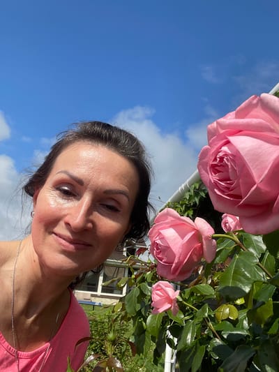 A woman is taking a selfie in front of a rose bush with pink roses. She is wearing a pink t-shirt and a silver necklace. The sky is blue with some white clouds. In the background, there is a house with windows and a garden. The image appears to be taken in the city of Solov'evka, Russia.