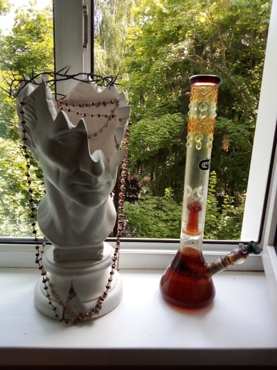 In a windowsill setting in посёлок дачного хозяйства «Архангельское», Russia, two objects are positioned against a backdrop of lush green trees. On the left, a white bust, resembling a classical sculpture, is adorned with a crown of thorns and a string of beads. The bust has a hollowed-out top. To the right of the bust, a clear glass bong, filled with a reddish-brown liquid, stands upright. The bong features decorative orange swirls near its top and a black circular logo. The scene is illuminated by natural light filtering through the trees.