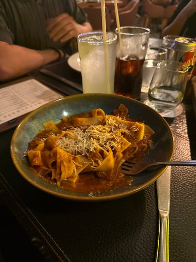 The scene depicts a dining experience within a dimly lit restaurant, presumably in San Agustín, Mexico. In the immediate foreground, a teal-rimmed ceramic bowl contains a serving of pappardelle-style pasta covered in a rich red meat sauce, generously topped with grated white cheese. A silver fork rests within the pasta, with a matching silver dinner knife positioned to the right on a dark, textured table surface or placemat.

To the left and behind the pasta dish, a menu with red lettering is partially visible and out of focus. Further back, on the left side of the frame, the arm and torso of an individual are visible, wearing a dark, possibly striped short-sleeved shirt. Their left arm, adorned with a dark wristband, rests on the table. Their hands are partially visible near a light-colored, frosty glass containing a yellow beverage with ice and two bamboo straws.

Adjacent to this drink is another tall clear glass holding a dark brown, ice-filled beverage, likely cola. Further to the right, a clear glass of water stands next to a partially visible silver Coca-Cola can. Another small, clear glass with a translucent liquid is also present on the table. The background is blurred, showing additional patrons or restaurant furniture, including chairs with reddish upholstery, illuminated by warm, ambient lighting. The overall environment suggests an evening meal in a casual dining establishment.