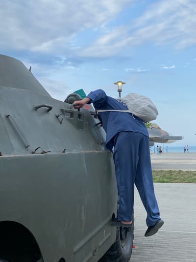 In Arablyar, Russia, a person in a blue jumpsuit and carrying a light-colored bag over their shoulder is climbing onto a green military vehicle. The person's left foot is on the vehicle's tire, and their right foot is on a step. The vehicle appears to be a BTR-60. In the background, there is a jet on display, a lamppost, a paved area, and a body of water. Several people are walking in the distance. The sky is cloudy.