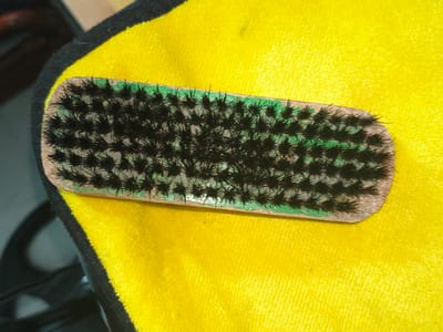 A close-up shot focuses on a small brush with black bristles, resting on a bright yellow, textured fabric. The brush has a wooden handle with a light brown finish. A thin band of green paint is visible along the edge of the handle where the bristles are embedded. The bristles are densely packed and stand upright, forming a textured surface. The yellow fabric appears soft and possibly plush, with a subtle, uniform texture. The overall composition is simple, with the brush as the primary subject against the vibrant backdrop. The lighting is bright, highlighting the details of the brush and the fabric. No people, text, or specific environmental cues are visible that would suggest a particular time of day, weather, or detailed location beyond the fabric and brush itself.