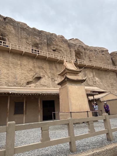 The image depicts a section of the Yulin Grottoes, a Buddhist cave complex located near Guazhou County, Gansu Province, China. A multi-tiered wooden structure is built against the rock face, providing access to cave entrances. The structure features ornate, traditional Chinese architectural elements like upturned eaves. A scaffolding-like structure climbs along the cliff. Two people are visible on an exterior platform. The environment is dominated by the light-colored rock of the cliff face and a gray sky.