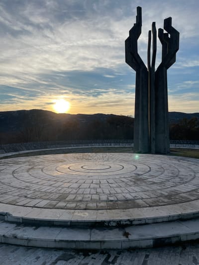 The image depicts an outdoor concrete monument situated in Barutana, Montenegro, at sunset. A large, multi-faceted concrete structure, approximately 10-15 meters in height, stands slightly right of center, characterized by dark grey, angular vertical elements that converge at its base and splay upwards, suggesting a flame or abstract hands. This monument is erected on a circular plaza paved with light-colored concrete pavers arranged in distinct concentric rings. The plaza foreground includes a two-tiered stepped edge. Beyond the plaza, a low, curved concrete wall defines the perimeter, followed by a grassy area and rolling hills densely covered with dark deciduous trees and shrubbery extending to the horizon. The sun, a prominent bright orb, is visible low on the horizon, positioned left of the monument, casting a warm, golden light across the scene. The sky above features scattered, wispy clouds against a gradient of blue and orange hues, indicative of late afternoon or early evening. No individuals, animals, or active subjects are present within the frame.