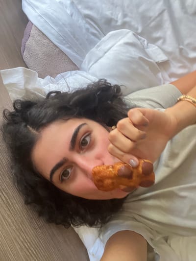 A young woman with dark, curly hair and light complexion lies on her back in an indoor setting within Orléans, France. She wears a light-colored t-shirt and a gold-toned bracelet on her left wrist. Her right hand, with light taupe nail polish, holds a golden-brown pastry resembling a pain au chocolat with dark chocolate pieces visible, positioned near her mouth. She looks directly at the camera with her dark eyes. The background consists of a light brown wooden floor or surface to her left, and white bedding with a textured light purple/beige blanket or bedspread to her right and behind her.