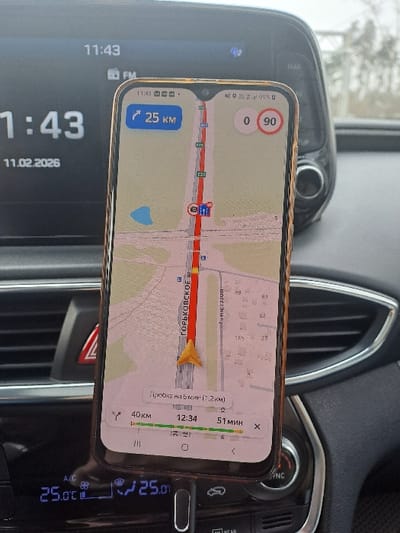The image depicts the interior of a vehicle, specifically focusing on a smartphone mounted vertically in a holder, displaying a navigation application. The scene is located in Obukhovo, Russia.
The smartphone screen shows a map centered on Gorkovskoye Shosse (ГОРЬКОВСКОЕ Ш.), with a yellow-orange arrow indicating the current vehicle position. A red line traces the route, and a traffic jam is indicated as "Пробка на 5 мин (1.2 км)" (Traffic jam for 5 min (1.2 km)). The navigation interface displays "25 KM" (likely distance to next turn or destination), "0 90" (possibly current speed and speed limit), total distance of "40 км," an estimated arrival time of "12:34," and remaining travel time of "51 МИН." The phone's status bar also shows "11:43."
In the background, the car's infotainment screen is visible, displaying "11:43" and the date "11.02.2026." A "FM" icon is also present. Below the smartphone, the vehicle's climate control panel shows a temperature setting of "25.0 °C" on both the left and right sides. A red hazard light button is partially visible to the left of the phone. The dashboard surfaces appear dark grey or black. The blurry background seen through the windshield suggests an outdoor environment with indistinct structures.