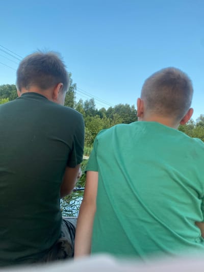 The image shows a close-up, rear view of two young boys from the shoulders up, sitting beside each other. Both boys have short, light brown hair and are wearing t-shirts, one dark green and the other a lighter shade of green. They appear to be outdoors, with a clear blue sky above and a treeline in the background. In the foreground, there is water with reeds, suggesting they are by a lake or river. The scene is calm and casual, and the boys seem to be engaged in a shared activity, possibly fishing, as a fishing rod is partially visible. The overall setting suggests a peaceful, natural environment, likely a recreational area in or near Afanasyevo, Russia.