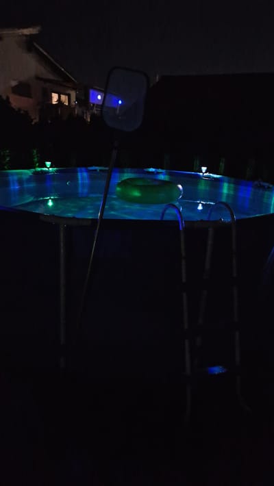 At night, an above-ground swimming pool is illuminated by blue and green underwater lights. A green inflatable ring floats in the pool, and a ladder with blue lights on its sides is attached to the pool. A pool skimmer is positioned over the pool, and a house with lights on is visible in the background. The scene is located in Ichenhausen, Germany.